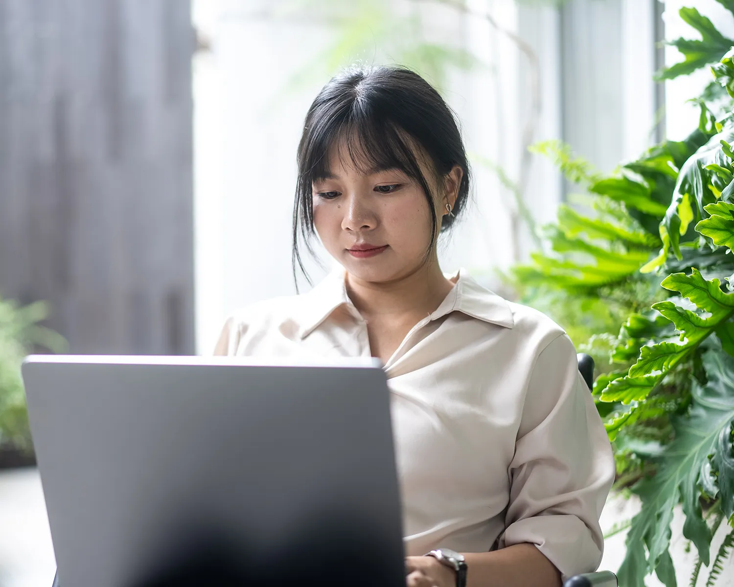A focused woman working on a laptop in a bright office setting, surrounded by large, vibrant indoor plants and natural light.