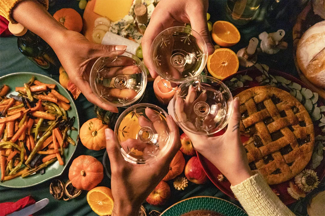 Hands raising glasses in a celebratory toast above a Thanksgiving dinner table filled with various dishes.