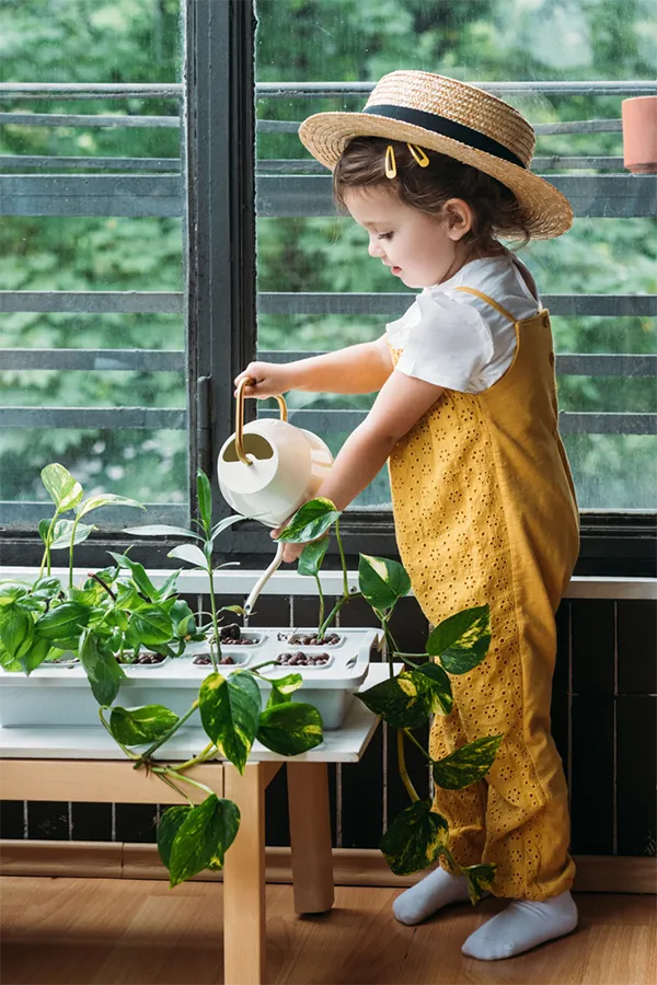 A young child in a straw hat and overalls waters a row of plants by a windowsill.