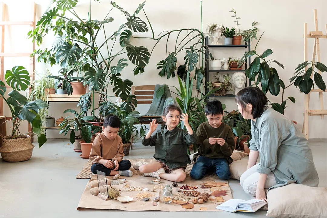 Four young children sit on a floor indoors with an adult, surrounded by numerous large potted plants and natural craft materials.
